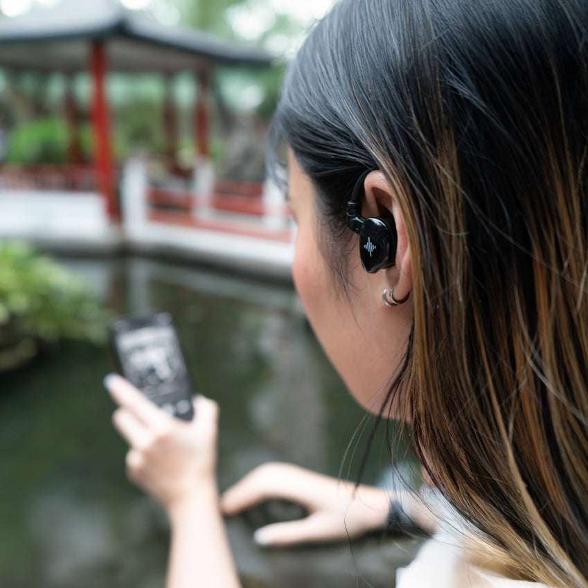 Person wearing a black in-ear earbud outdoors while holding a smartphone