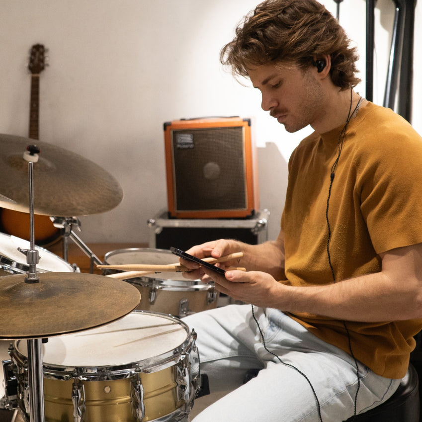 Man wearing in-ear monitors while seated at a drum kit and holding a smartphone
