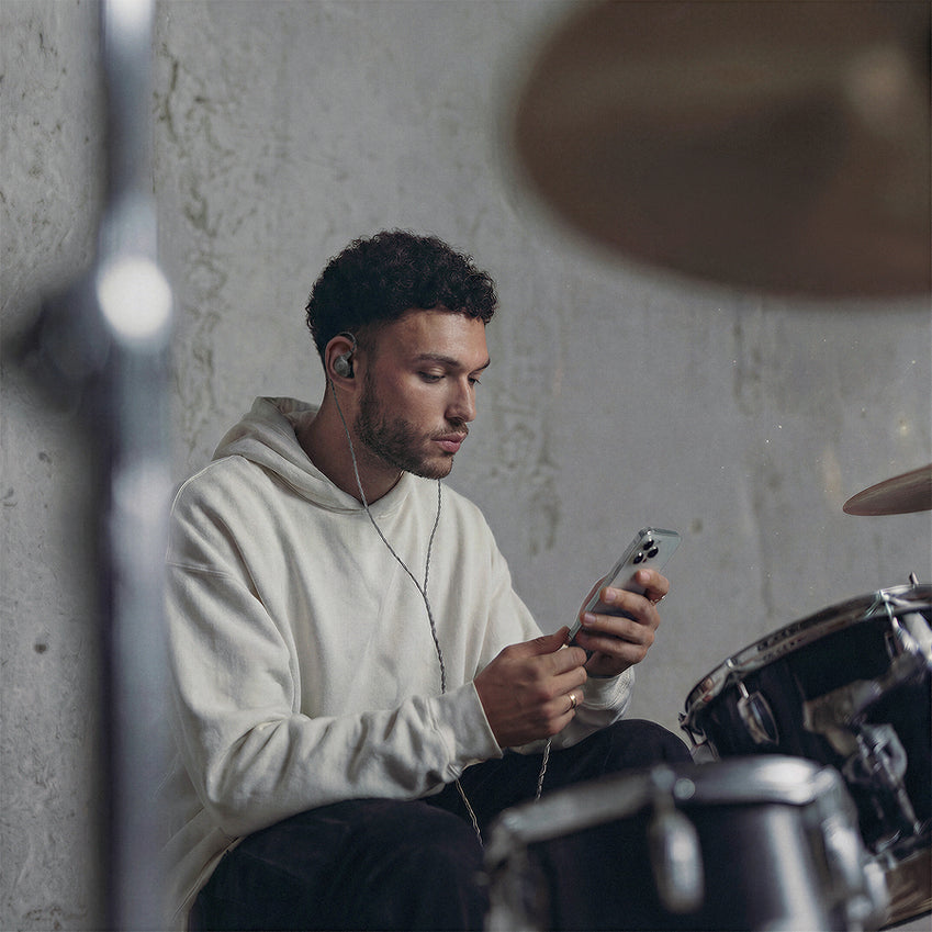 Musician wearing an in-ear monitor looks at a phone beside a drum kit